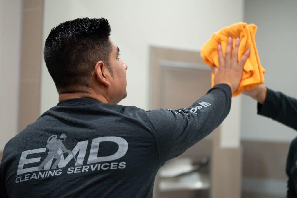 A person wearing an EMD Cleaning Services shirt uses a yellow cloth to clean a mirror in a restroom, demonstrating the thoroughness of their commercial cleaning standards.