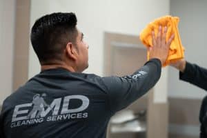 A person wearing an EMD Cleaning Services shirt uses a yellow cloth to clean a mirror in a restroom, demonstrating the thoroughness of their commercial cleaning standards.