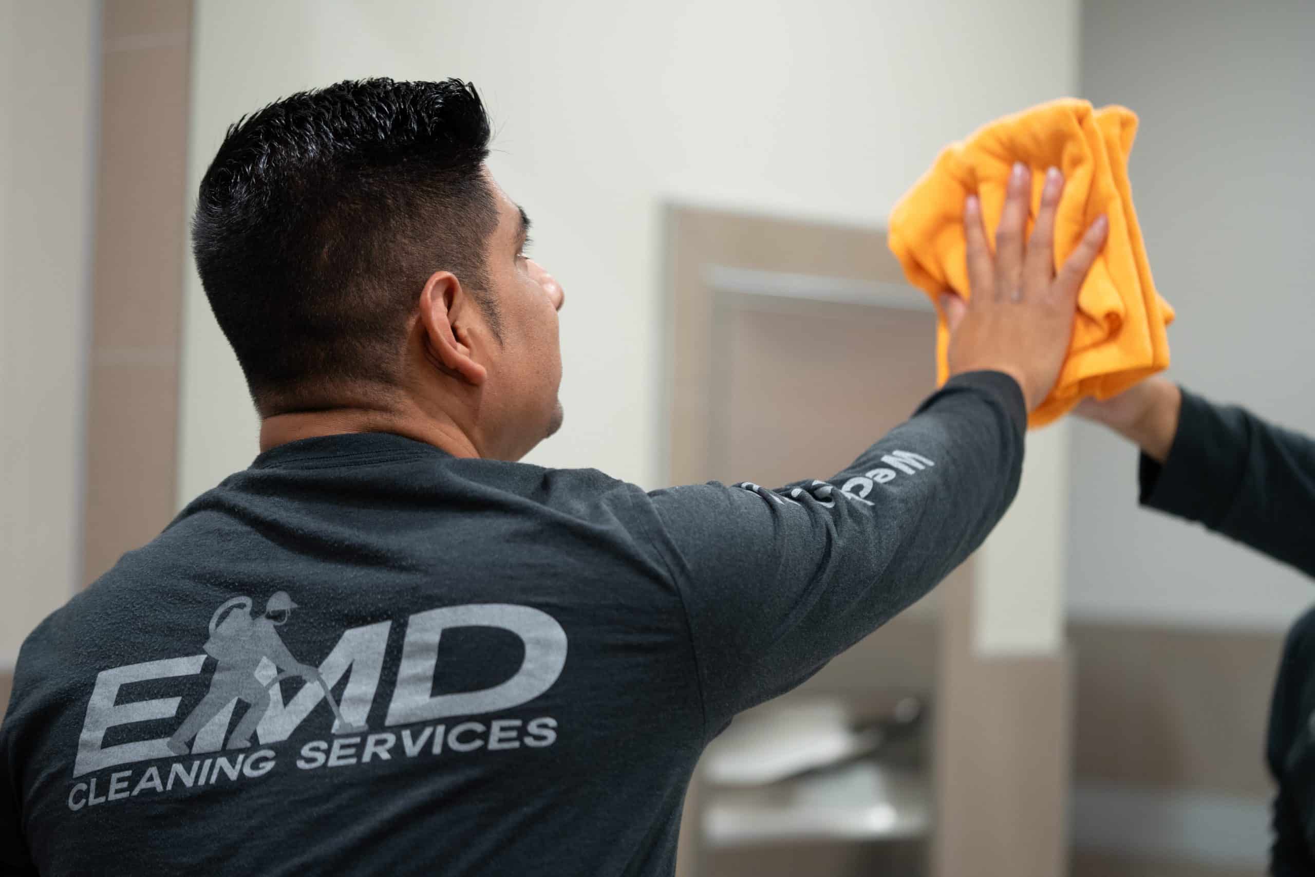 A person wearing an EMD Cleaning Services shirt uses a yellow cloth to clean a mirror in a restroom, demonstrating the thoroughness of their commercial cleaning standards.