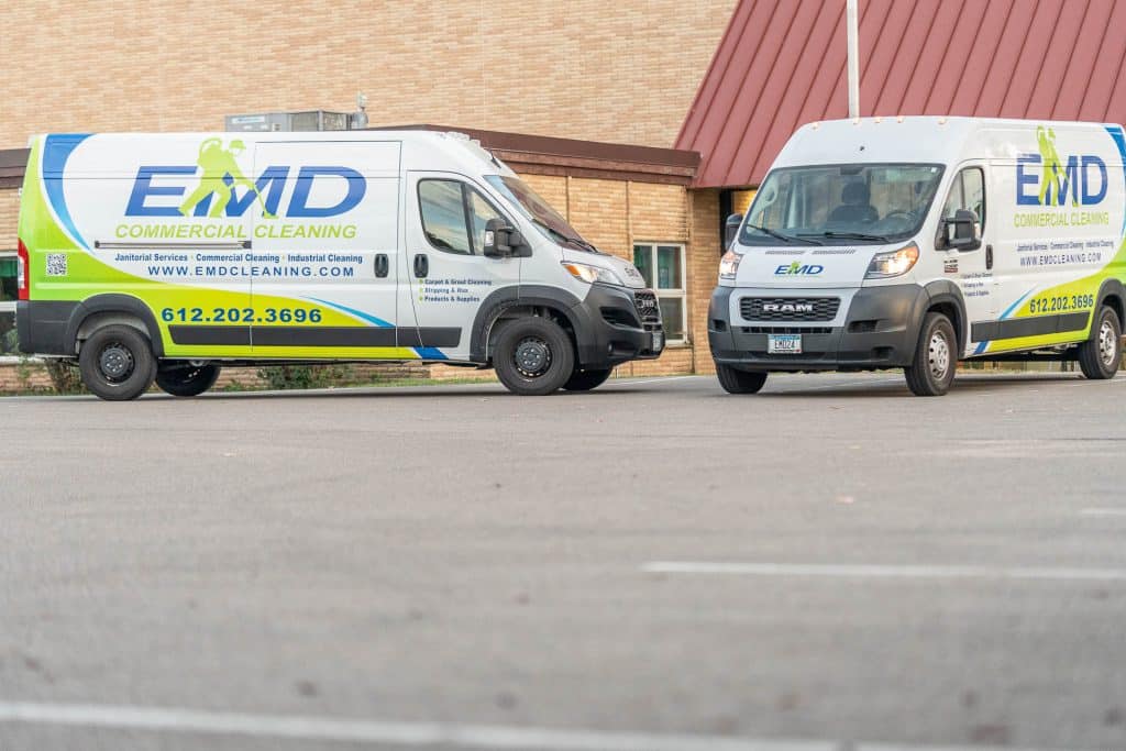 Two white vans with EMD Commercial Cleaning logos and contact information are parked in a lot in front of a building with tan brick walls and a red roof, ready to provide expert office cleaning and medical office cleaning services.