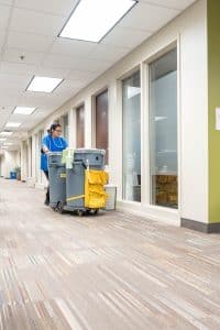 A janitor pushes a cleaning cart down a brightly lit office hallway with beige walls, large windows, and patterned carpet, showcasing the professionalism of commercial cleaning services.